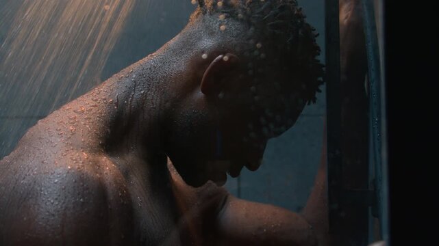 Tired depressed African American muscular man hitting wall under falling shower water sad male stressed guy thinking about health problem trouble anxiety pain crisis washing body inside dark bathroom