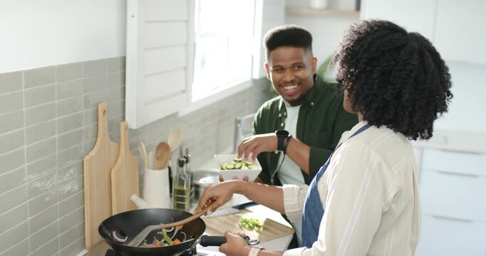African American couple at counter, man bringing bowl, woman stirring wok, man adding zucchini