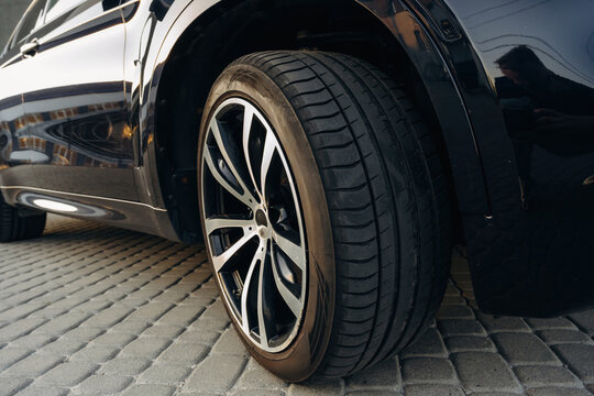Close-up view of a black alloy wheel mounted on a vehicle, showcasing intricate design and tread pattern on a cobblestone surface