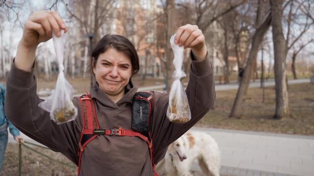 Woman wearing brown hoodie and red harness holds up plastic bags filled with dog waste in outdoor park setting. white long haired dog is visible behind her walking on path as another woman