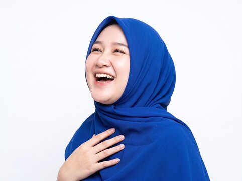Young Muslim woman dressed in vibrant blue hijab laughing joyfully with hand on chest against a clean white background