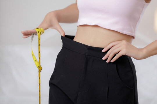 woman showing loose pants at waist while holding measuring tape, representing weight loss, body measurement, healthy lifestyle, fitness progress, diet goals, wellness transformation.