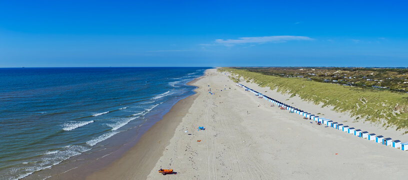 Aerial view of the sandy beach and dunes at Texel with rows of beach huts along the coastline under a clear blue sky in De Koog, North Holland, Netherlands.