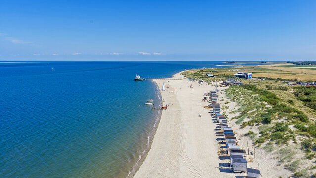 Aerial view of the sandy beach at Texel with a row of colorful beach huts nestled against the dunes, calm blue sea, and a ferry to Vlieland on Texel island, North Holland, Netherlands.