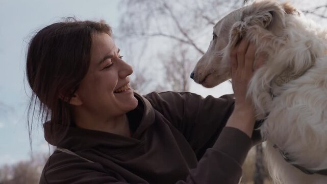 Woman interacts affectionately with large white dog outdoors holding and petting it. scene features natural daylight in open area with bare trees visible in background. framing emphasizes