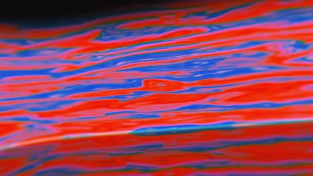 Horizontal iridescent oil slick surface with shifting red and blue fluid ripples in a dynamic psychedelic 3D animation