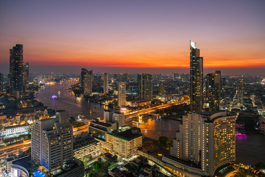 Aerial view of Chao Phraya River winding through the illuminated Bangkok skyline at sunset with modern skyscrapers and glowing city lights in Bangkok, Thailand.