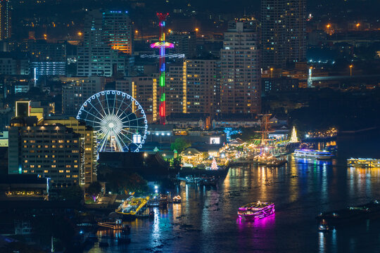 Aerial view of Chao Phraya River at night with the illuminated Ferris wheel at Asiatique, river boats, and city skyline in Bangkok, Thailand.