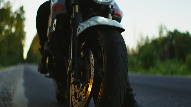 Powerful motorcycle riding on empty forest road at sunset, close-up details of bright headlight, spinning front wheel and dynamic motion of solo biker