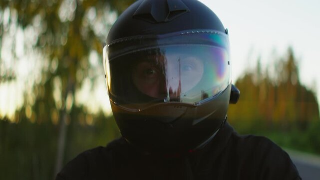 Motorcyclist wearing black full-face helmet with reflective visor riding through scenic forest road at sunset, closeup portrait capturing adventure and focus