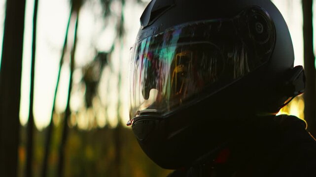 Motorcyclist wearing a reflective visor helmet rides through a tranquil forest at golden hour as warm sunset light glows on the protective gear and surrounding trees