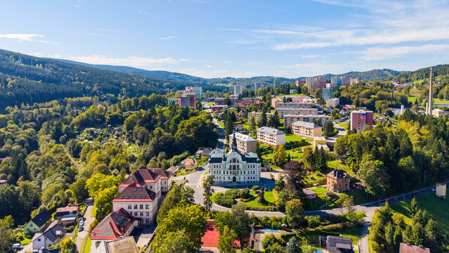 Aerial view of the historic town hall surrounded by residential buildings, lush green forests, and rolling hills in Tanvald, Liberec Region, Czechia.