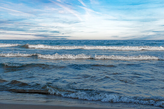 Gentle ocean waves gently lap against a sandy shore under a bright blue sky filled with wispy clouds.