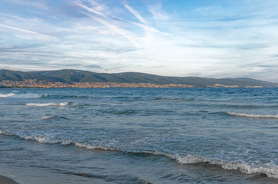 Gentle waves lap against the sandy shore with mountains and a coastal city visible under a soft blue sky across the water.