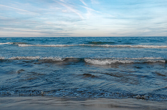 Gentle blue sea waves roll onto a sandy beach under a light blue sky with faint white clouds.