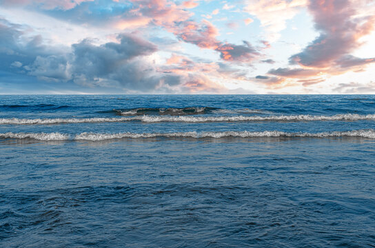 Gentle waves roll across a calm blue sea under a colorful sunset sky filled with pink and gray clouds.
