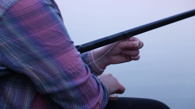 Close up of hands baiting a hook near a fishing rod against a calm gray lake. Fingers pinch and thread the bait carefully, moving slowly while preparing the tackle for casting.