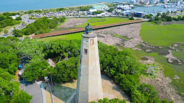 Bald Head Island, United States - 17 June 2023: Aerial view of Old Baldy Lighthouse and Smith Island Museum surrounded by lush greenery, marshlands, and a wooden boardwalk.