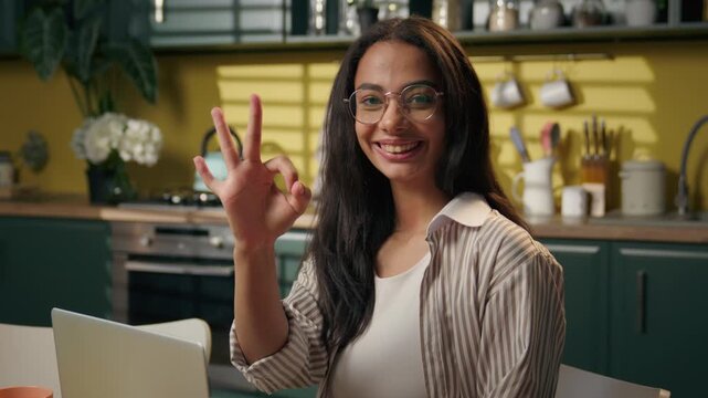 Smiling satisfied Latina American teen girl at home with laptop showing ok sign happy Hispanic high school student businesswoman looking at camera excellent online study positive freelancer at kitchen
