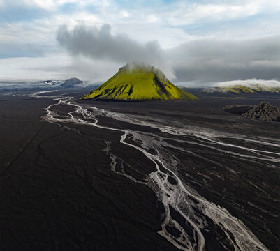Aerial view of Maelifell volcano covered in vibrant green moss, surrounded by vast black volcanic sand plains and intricate glacial river channels in Skaftarhreppur, Iceland.