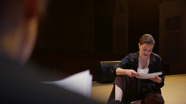 Focused female actor rehearsing lines on dimly lit theater stage during dramatic performance practice, seated on floor with script pages under warm spotlight