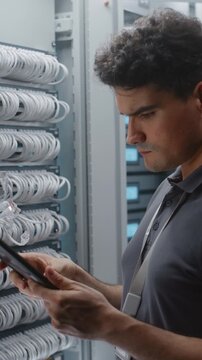 Male IT Engineer Checks Rack Connections While Consulting Tablet, Verifying Physical Cabling Against Network Records During Structured Infrastructure Inspection. Maintains Data Center Infrastructure