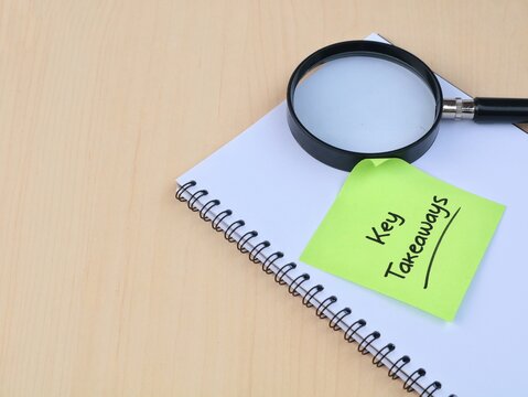Green adhesive note with handwritten Key Takeaways text placed on a spiral book with a magnifying glass on a wooden desk. Top view concept for business research, analysis, and key findings