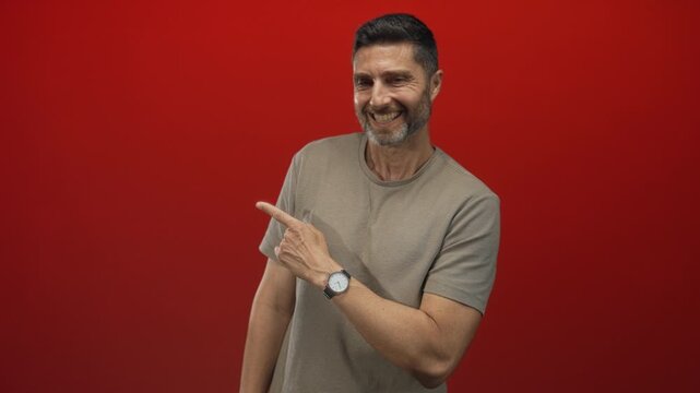 Man pointing finger in studio against a red backdrop, smiling and wearing a watch and tshirt; friendly confidence.