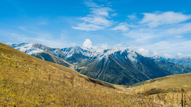 Landscape photograph of majestic snow-capped mountain range and golden autumn meadow viewed from Chkhutnieri pass in Svaneti Georgia.