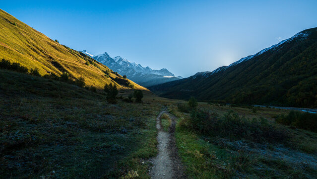 Landscape photograph of dirt hiking trail leading through mountain valley toward snow capped peaks under clear blue sky near Adishi in Svaneti Georgia.