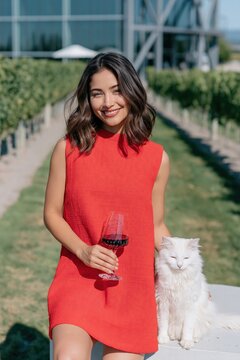 Woman in red dress holding a glass of red wine beside a white cat at a vineyard terrace for winery background and lifestyle banner design
