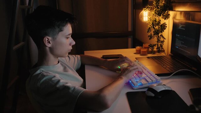 Teenage boy typing on an RGB keyboard and coding on a laptop at a desk in a dimly lit room at night.