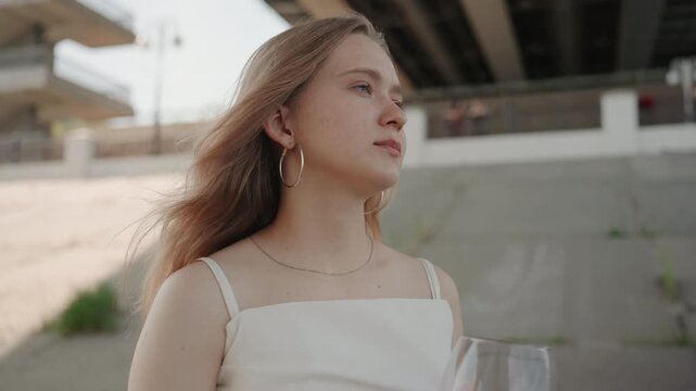 Young woman with long hair wears white dress and necklace holding or drinking from glass of red liquid outdoors under overpass. Her windblown hair suggests movement while background features
