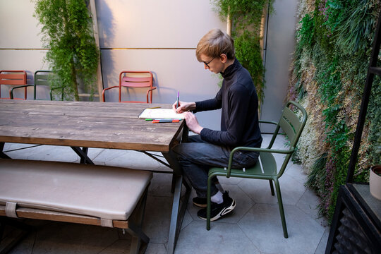 A young boy wearing glasses drawing while sitting at wooden table