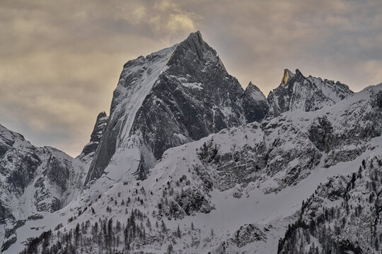 View of the snow-covered peaks of the Engadin mountains in the Swiss Alps, featuring rugged rocky crags and pine trees under a cloudy winter sky in Grisons, Switzerland.