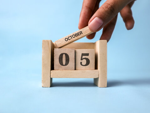 A woman's hand holds a wooden block with October written on it on a wooden block calendar for October 5th against a light blue background.