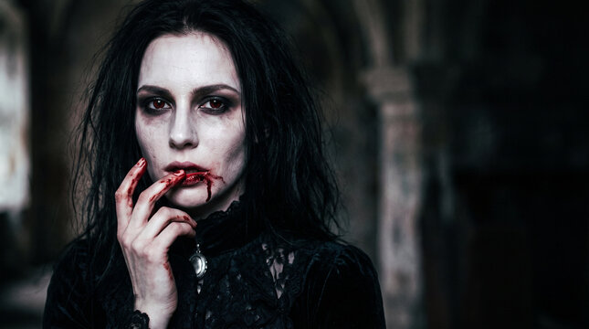 Cropped portrait of an eerie female vampire with pale skin, red eyes and blood-stained lips touching her mouth, wearing a black gothic dress in a dark Halloween horror setting.