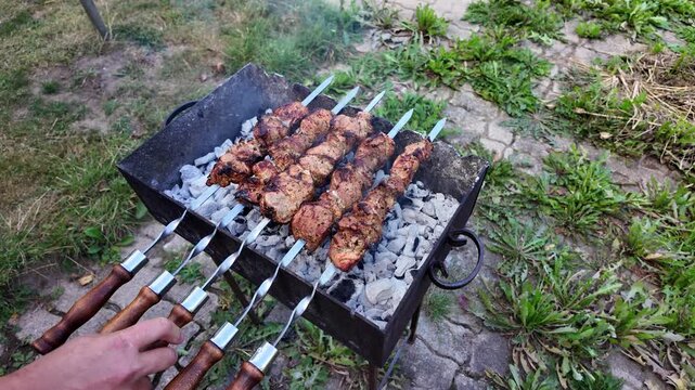 Human hands turning steel skewers with juicy chunks of grilling meat over hot coals on an outdoor barbecue.