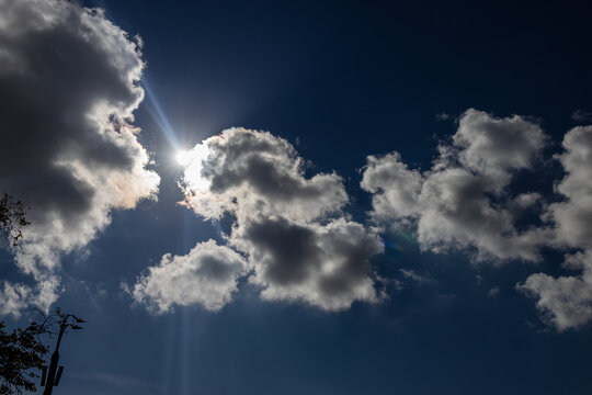 Bright sun rays breaking through fluffy cumulus clouds in a dark blue sky with bright backlit cloud edges.