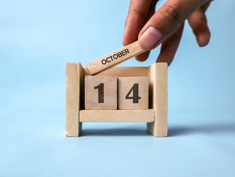 A woman's hand holds a wooden block with October written on it on a wooden block calendar for October 14th against a light blue background.