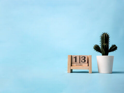 A wooden block calendar for October 13th with a plastic cactus plant on the side on a light blue background.