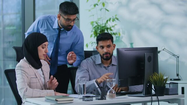 Arabian business team partnership teamwork in office three Indian workers men with woman in hijab communicate looking at computer screen analyze documents work data discussing job strategy together