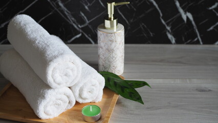 Luxury spa wellness still life with rolled white towels, elegant soap dispenser and green candle on a wooden tray against a dark marble background, creating a calm relaxation atmosphere. © kanjana