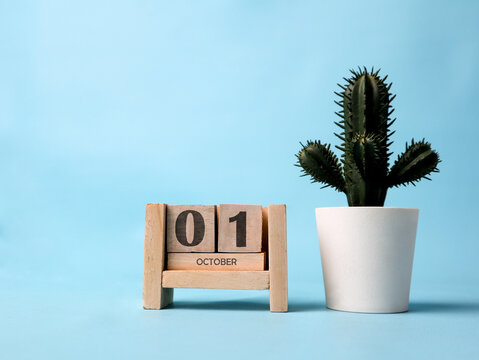 A wooden block calendar for October 1st with a plastic cactus plant on the side on a light blue background.
