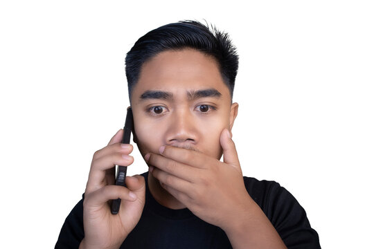 Shocked Indonesian man wearing a black t-shirt talking on a mobile phone while covering his mouth with his hand and looking wide-eyed against a white background.