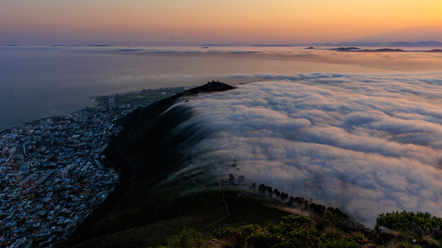 View of Lion's Head with a thick layer of clouds rolling over the mountain at sunset, overlooking the city lights and the coastline in Cape Town, Western Cape, South Africa.
