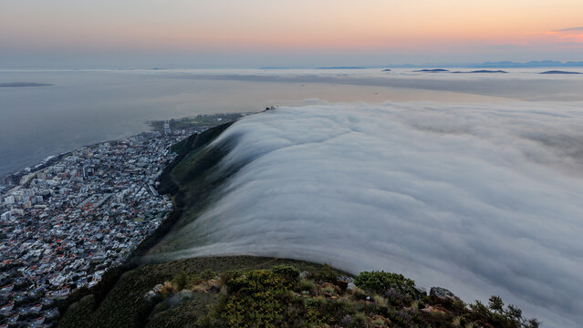 View of Lion's Head with a thick cloud bank rolling over the mountain slopes at sunset, overlooking the city and the Atlantic Ocean in Cape Town, Western Cape, South Africa.