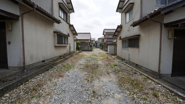 戸建て住宅の間に細長く続く砂利と土の狭小な住宅街空き地、側溝と両側の白い外壁の景観