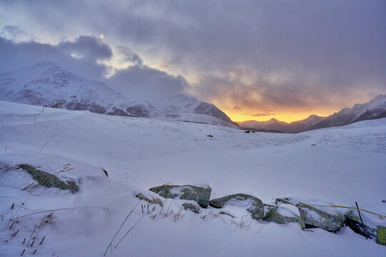 View of the snow-covered Valchiavenna mountains at sunset with a stone wall in the foreground under a dramatic cloudy sky in Madesimo, Lombardy, Italy.