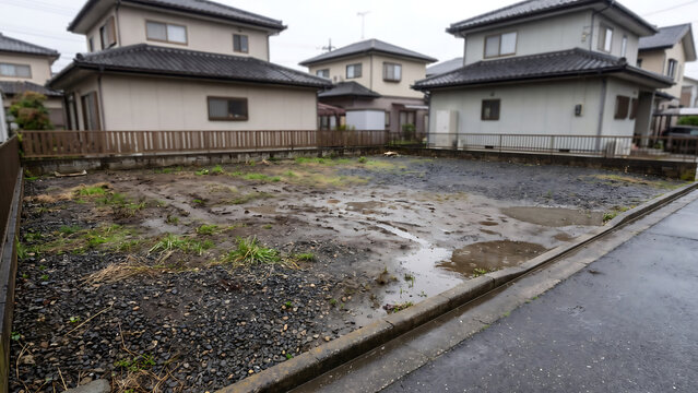 浅い水たまりと濡れた砂利が見える住宅街の空き地と戸建て住宅、生活道路とコンクリート縁石の景観